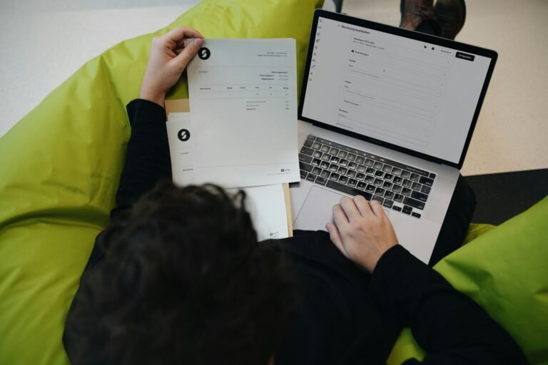 Freelancer learning how to keep track of invoices by comparing a printed invoice against invoicing software on a laptop