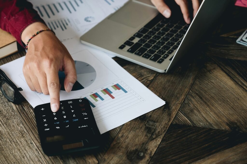 Freelancer reviewing printed financial charts and using a calculator alongside a laptop on a wooden desk