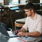 Freelancer learning how to manage freelance projects with clients on a laptop at a café table with a notebook and phone