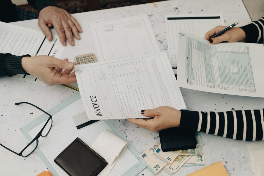 Two people reviewing and exchanging a printed invoice alongside tax forms, a calculator, and euro banknotes on a desk