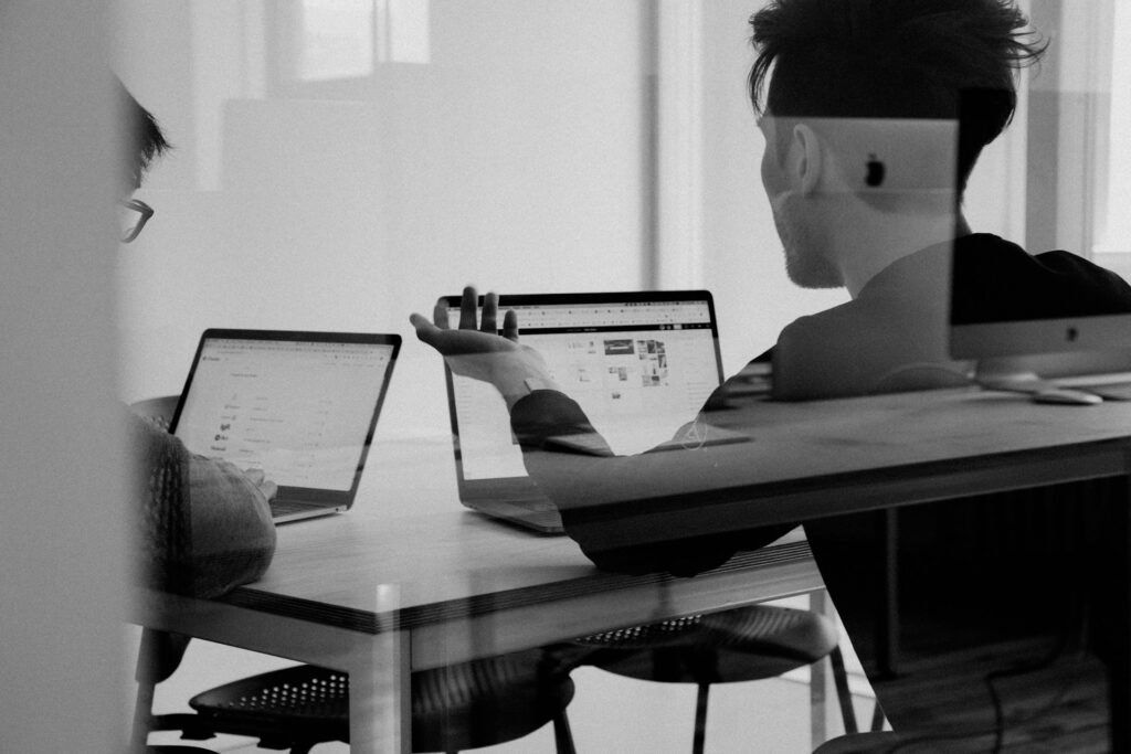 A client and a freelancer sit in a meeting room on laptops while talking about an ongoing project