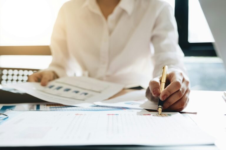 Freelancer reviewing printed financial reports and charts with a pen and bookkeeper software at an office desk.