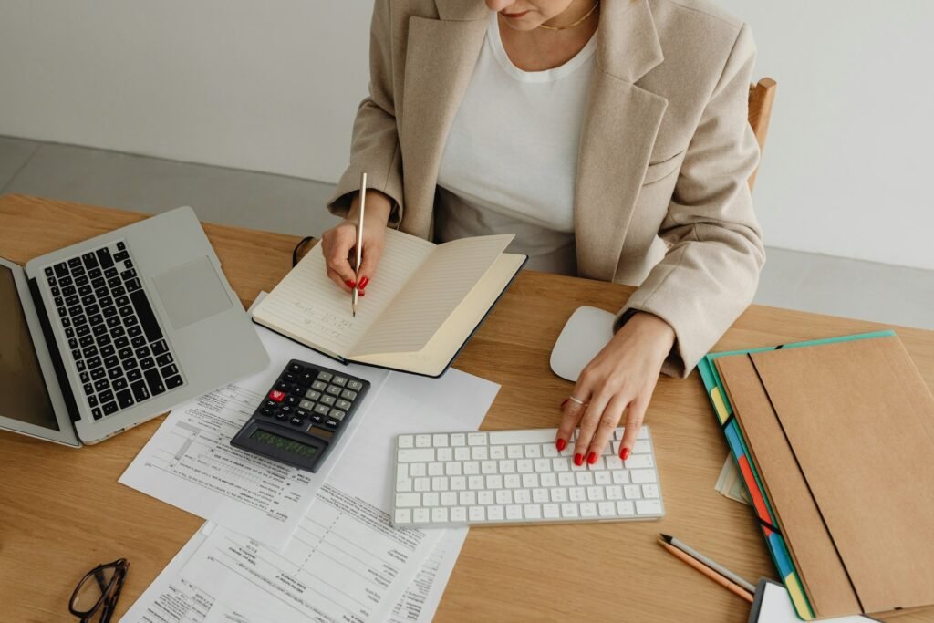 Freelancer writing notes in a journal while using a calculator and keyboard to manage business finances at a desk