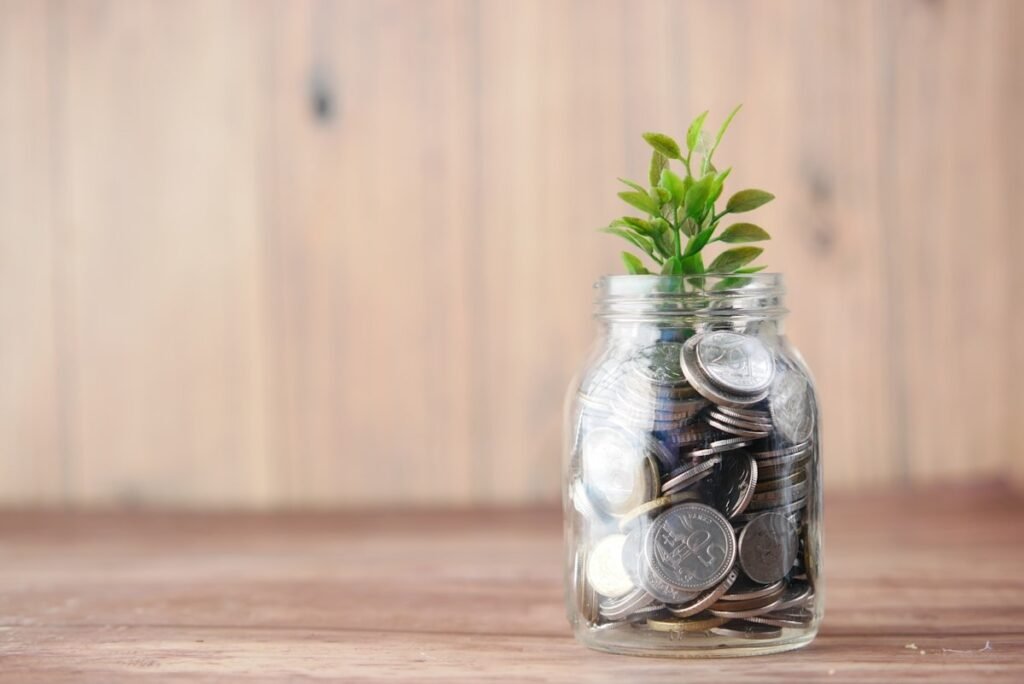 A glass jar filled with coins with a small green plant growing from the top, symbolizing a growing savings fund.