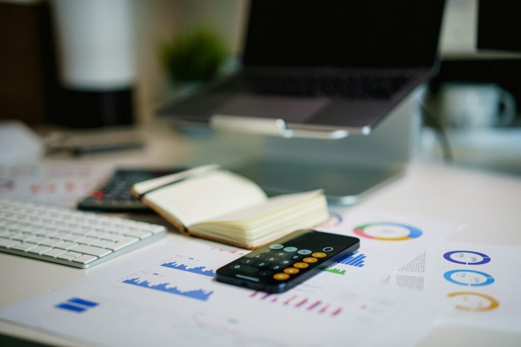 Smartphone calculator resting on printed financial charts beside a notebook and laptop on a desk 