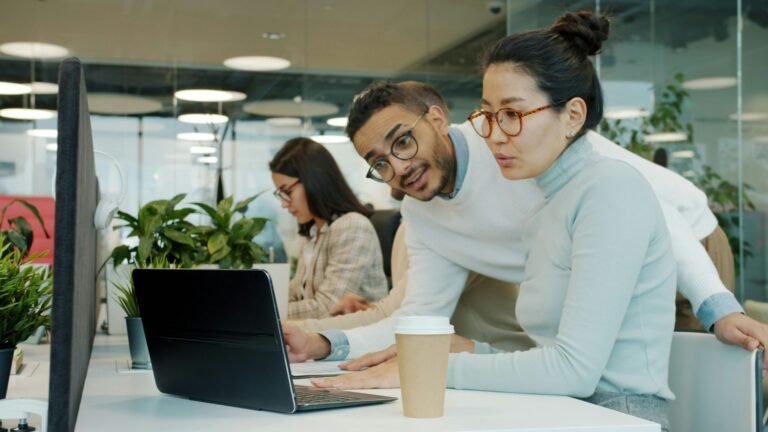 Two colleagues sitting together in an office, talking and reviewing work on a laptop screen, after reflecting on how to assign tasks to freelancers.