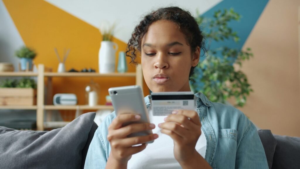 A woman sits on a couch, focused on a smartphone and holding a credit card. The background shows a shelf with plants and geometric wall decor.