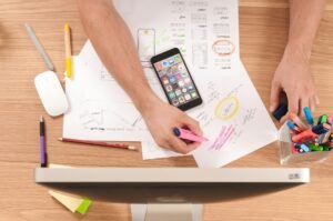 Best Freelancer Management Tools for Multi-Project Workflows: freelancer planning multi-project workflows with notes, app wireframe sketches, phone, and pens.