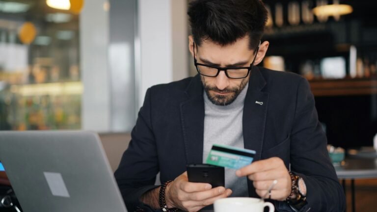 Freelancer sitting at a table holding a credit card while using one of the best tools for comparing bank accounts on his phone.