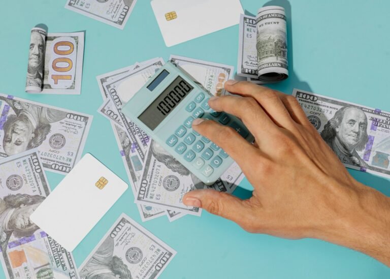 Hand using a calculator on a desk scattered with US hundred-dollar bills and bank cards, illustrating a freelancer weighing up cash flow forecasting software