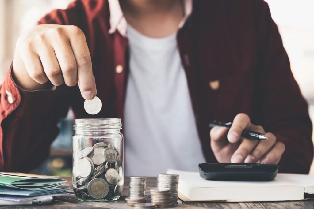 Hand placing a coin into a glass jar beside a calculator, illustrating a freelancer's monthly tax set-aside