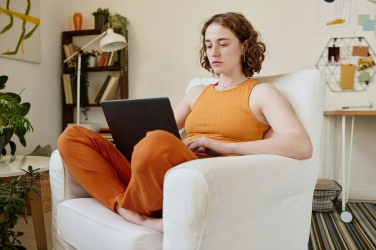 A female freelancer in orange sits on her laptop in a chair working out her freelance tax deductions for the year.