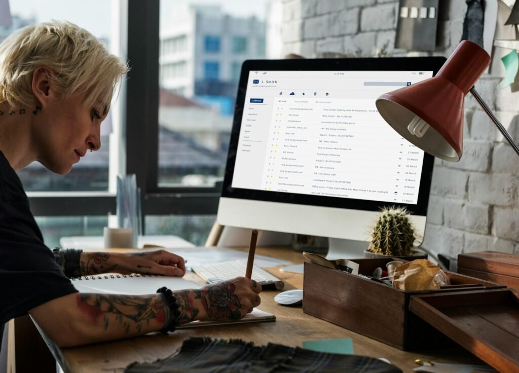 Freelancer taking notes at a desk beside a monitor displaying an email inbox