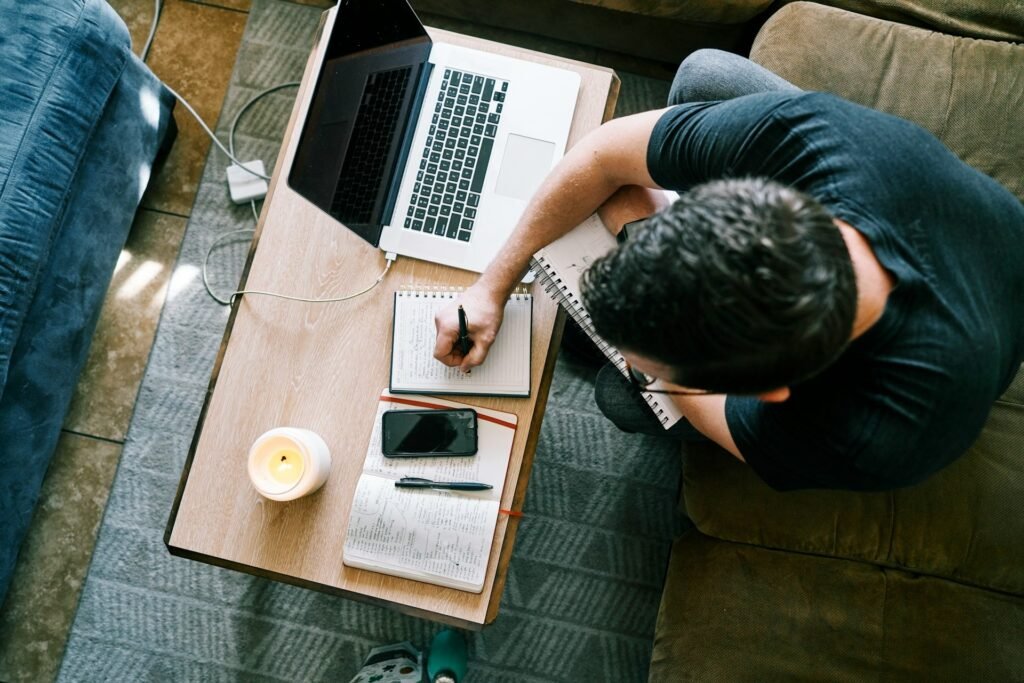 A top-down view of a freelancer working at his desk writing notes, with a laptop and computer open in front of him