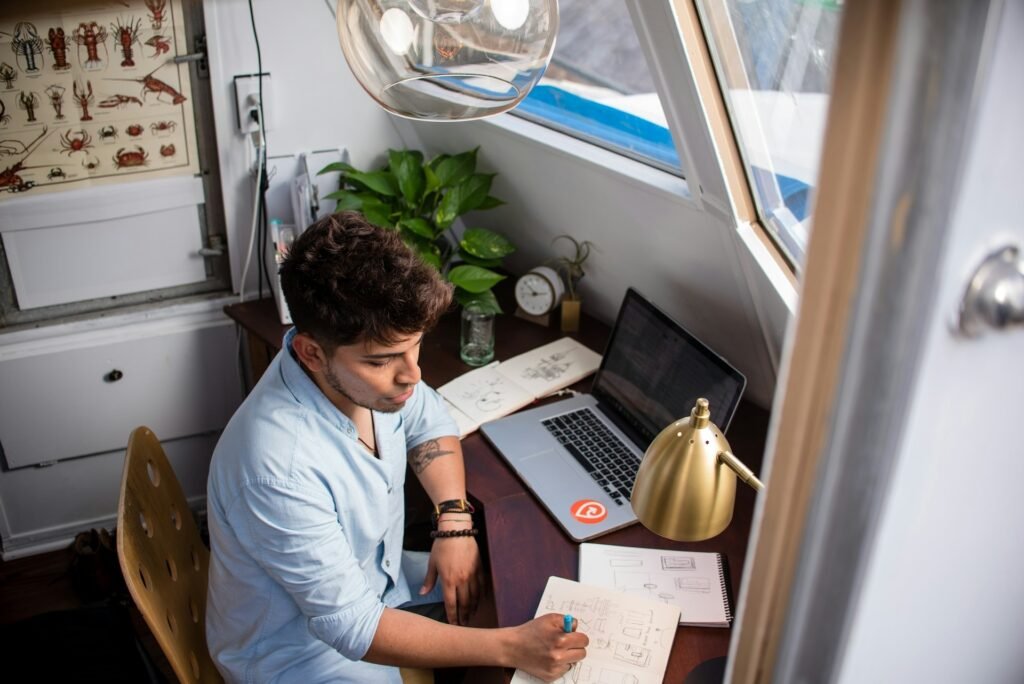A freelancer works on a project at his desk with a clock next to his laptop for tracking the time