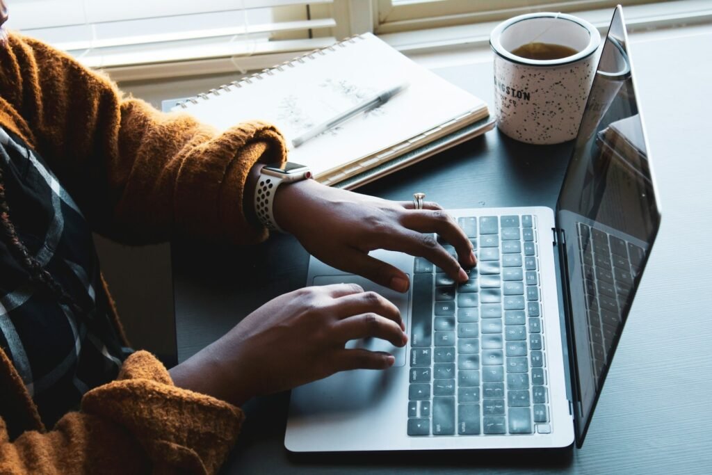 Hands typing on a laptop beside a closed notebook and a mug of tea