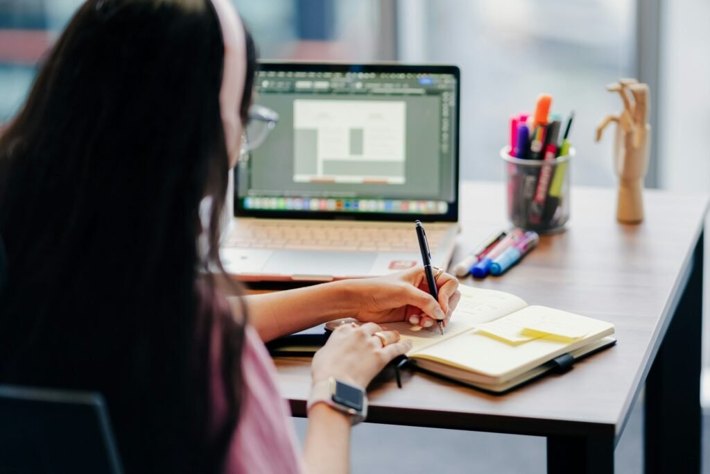 Notes being written in a lined notebook with sticky tabs beside a laptop