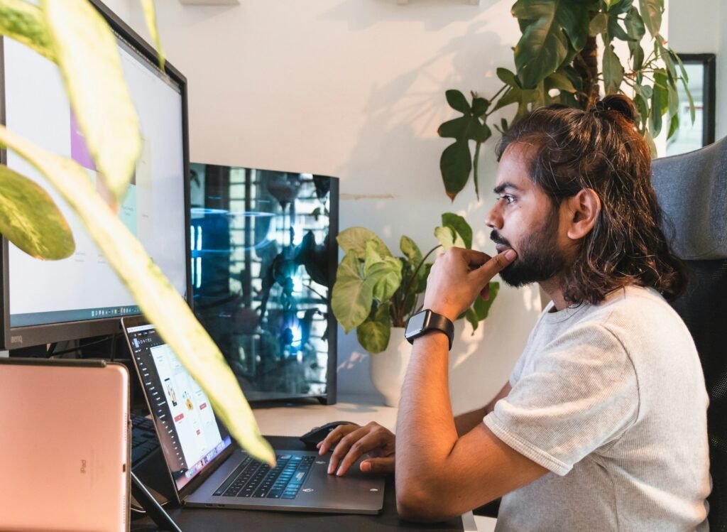 Freelancer pausing at a two-screen desk setup, thinking through the work in front of him