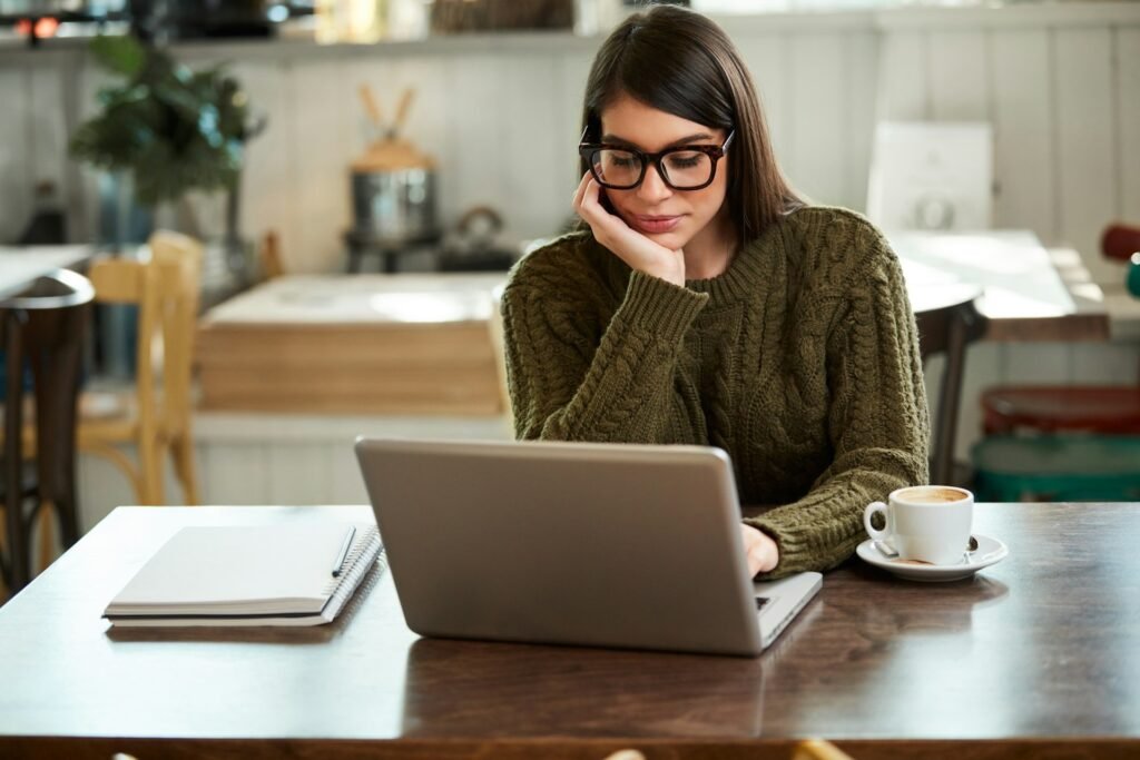 Freelancer reading at a laptop in a cafe, chin resting on her hand, notebook and coffee beside her