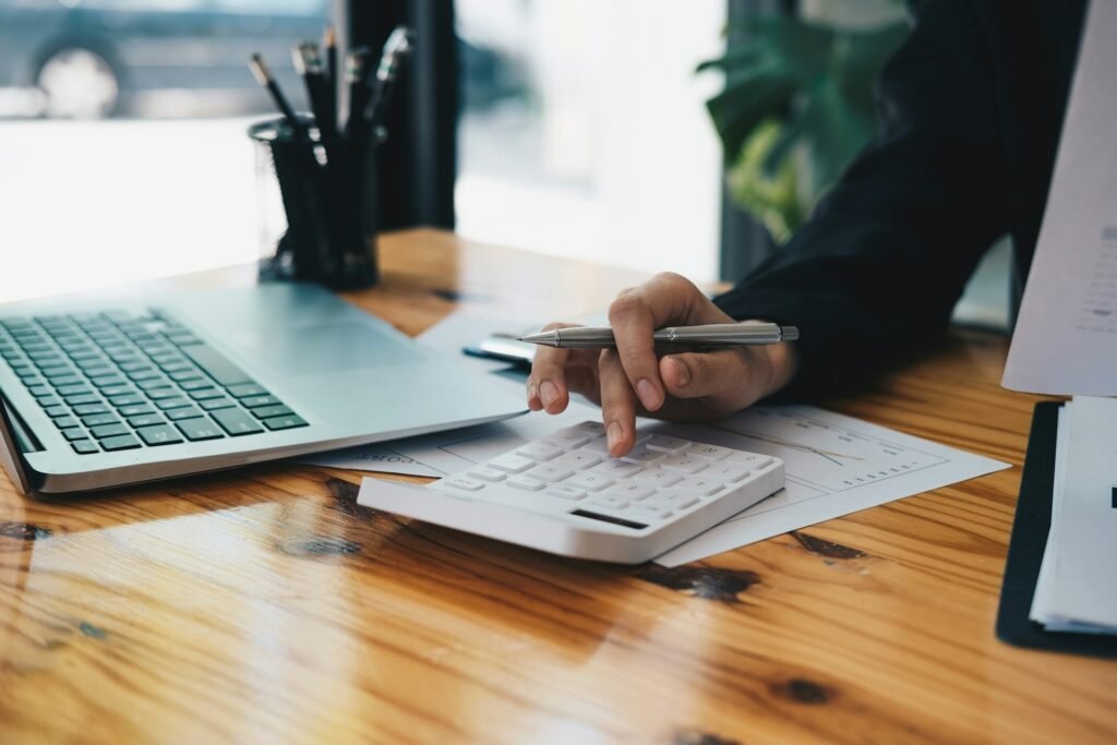 Freelancer using a calculator beside a laptop and printed financial paperwork