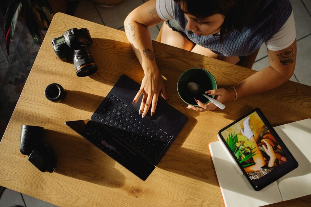 Freelancer working at a laptop with a camera and tablet on the desk, eating lunch one-handed