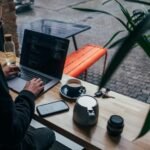 Freelancer working on a laptop at a cafe window bench, phone face-up on the table beside him