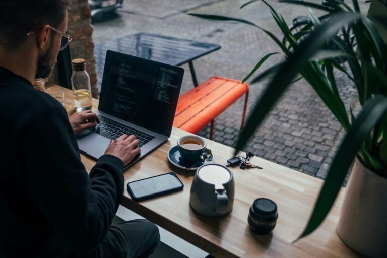 Freelancer working on a laptop at a cafe window bench, phone face-up on the table beside him