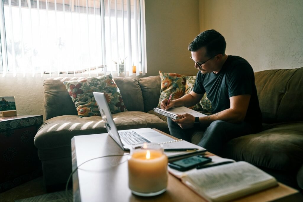 Freelancer working from a sofa, writing in a spiral pad with a laptop open on the coffee table
